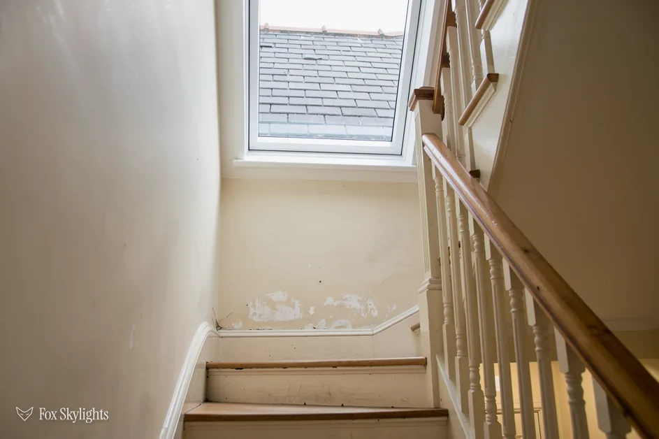 Roof window above a staircase with a timber banister and scuffed wall