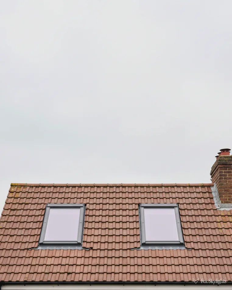 Two roof windows side by side in a red tiled roof below a chimney