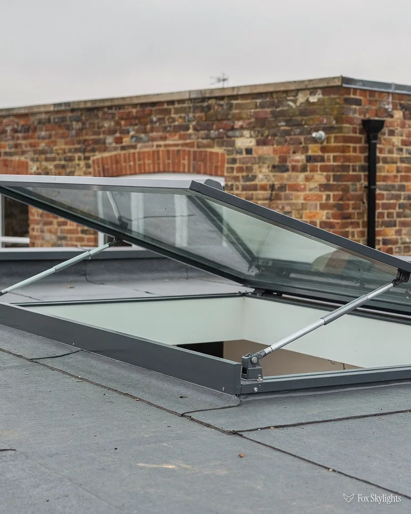 Opening rooflight propped open on a flat roof beside a brick wall