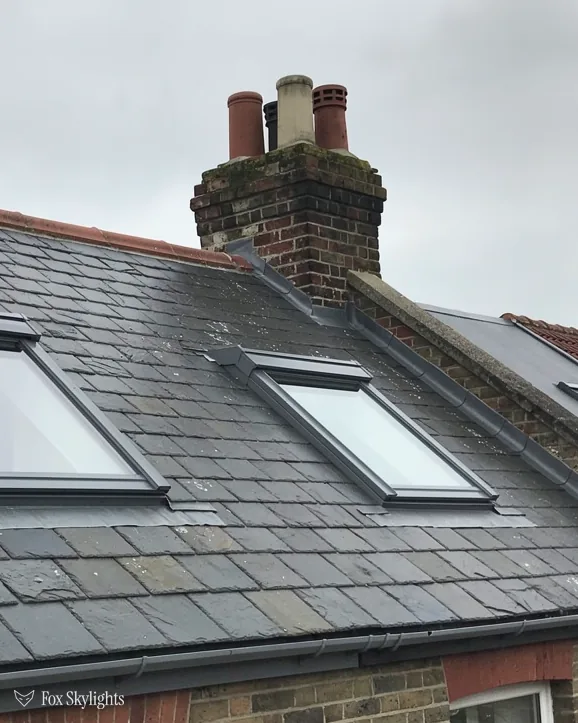 Pair of roof windows set in a slate roof beneath a rendered chimney stack